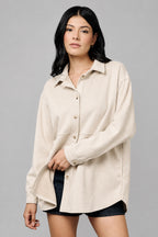 A woman wearing an oversized, long-sleeve button-up shirt shacket in an ecru color, paired with black shorts, against a textured neutral studio backdrop.