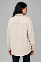 A woman shown from the back wearing an ecru color, long-sleeve collared shacket with dark pants, standing against a textured beige wall.