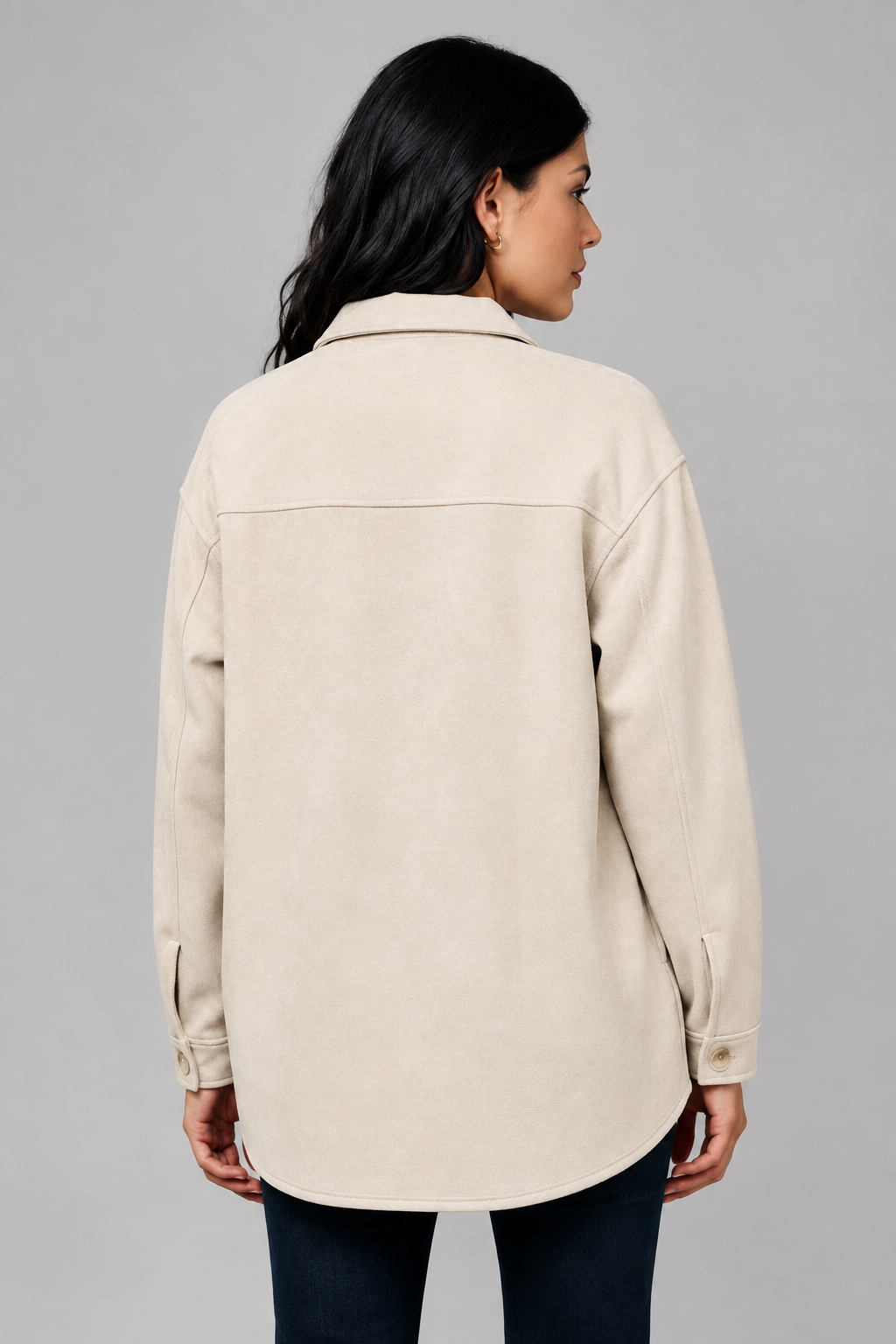 A woman shown from the back wearing an ecru color, long-sleeve collared shacket with dark pants, standing against a textured beige wall.