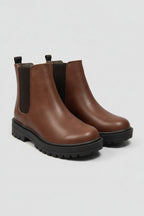 A pair of brown leather Chelsea boots with black elastic side panels and chunky black lug soles, shown on a light gray background.