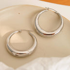 Two polished silver-plated hoop earrings with a thick, rounded crescent shape, resting on a light beige ceramic dish.