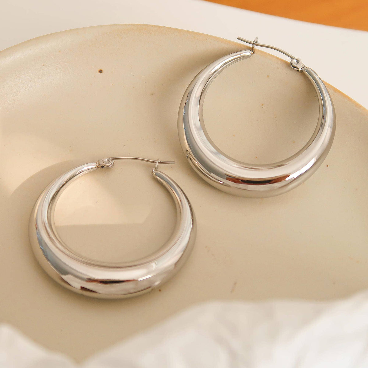 Two polished silver-plated hoop earrings with a thick, rounded crescent shape, resting on a light beige ceramic dish.