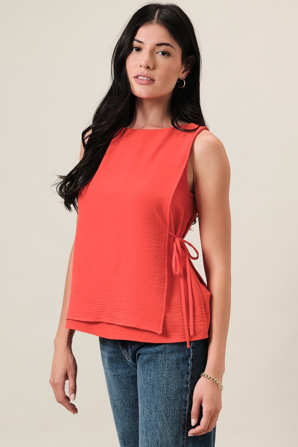 Woman wearing an orange sleeveless layered top and blue jeans on a beige background