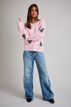 Woman wearing a pink sweater with green Christmas tree patterns and blue jeans on a plain background