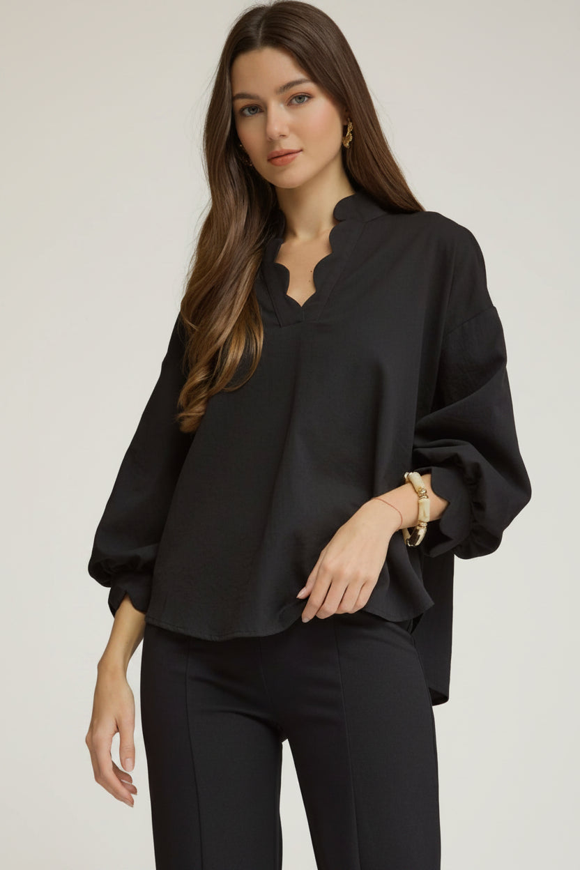 A woman modeling a black puff sleeve blouse with a scalloped V-neckline and cuffs, paired with black pants, against a neutral gray studio background.