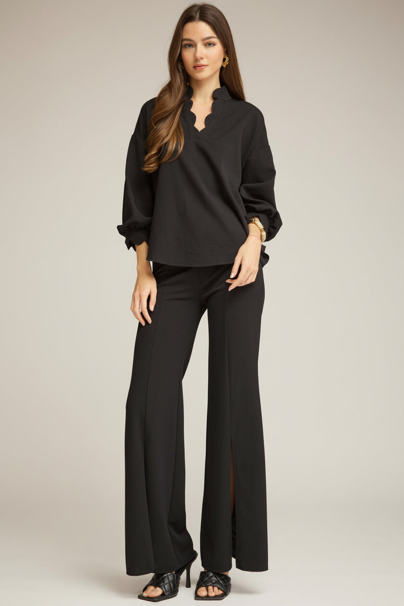 A woman modeling a black puff sleeve blouse with a split neckline and black wide-leg trousers with front slits, styled with black heeled sandals against a neutral gray studio background.