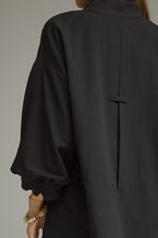 Close-up back view of a woman wearing a black blouse with puff sleeves and a pleat detail with tab accent against a plain light gray background.
