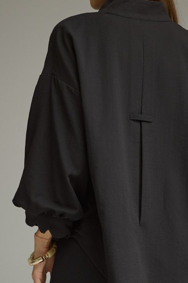 Close-up back view of a woman wearing a black blouse with puff sleeves and a pleat detail with tab accent against a plain light gray background.