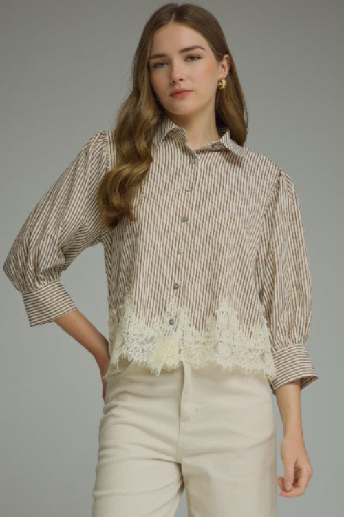 A woman modeling a brown and cream vertically striped button-up blouse with cream lace hem, paired with cream-colored pants, against a solid gray studio background.