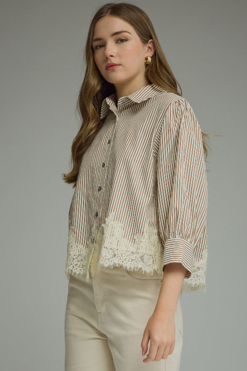 A woman modeling a brown and cream vertically striped button-up blouse with cream lace hem, paired with cream-colored pants, against a solid gray studio background.