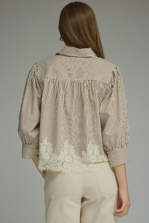 Back view of a woman modeling a brown and cream vertically striped button-up blouse with cream lace hem, paired with cream-colored pants, against a solid gray studio background.