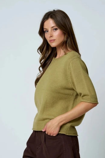 A woman wearing a short-sleeve olive knit top and dark brown shorts, photographed against a light gray studio background.