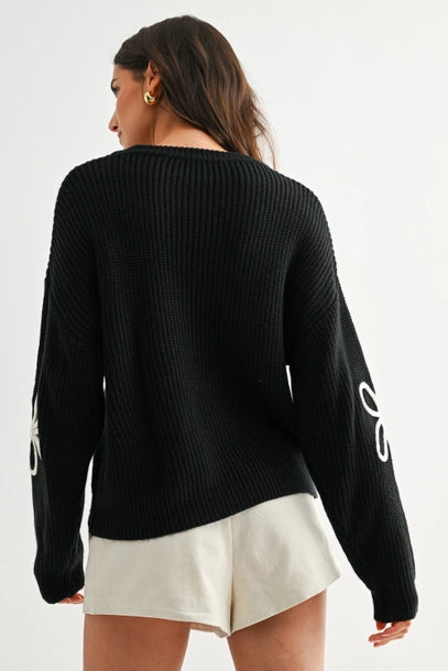 Back view of a woman wearing a black sweater with white daisy loop appliques on a white background