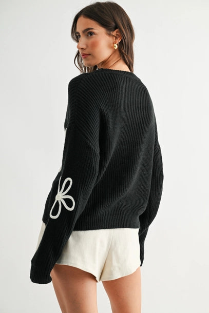 Side view of a woman wearing a black sweater with white daisy loop appliques on a white background