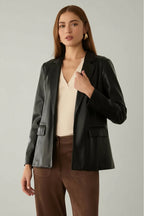 Woman wearing a black faux leather jacket over a beige top and brown pants in a neutral setting.