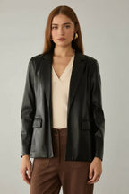 Woman wearing a black faux leather jacket over a beige top and brown pants in a neutral setting.