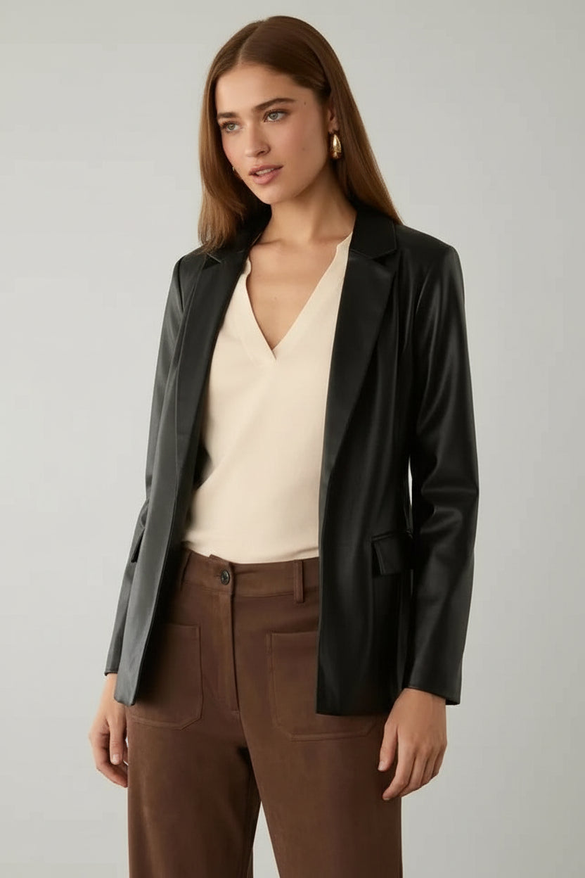 Woman wearing a black faux leather jacket over a beige top and brown pants in a neutral setting.
