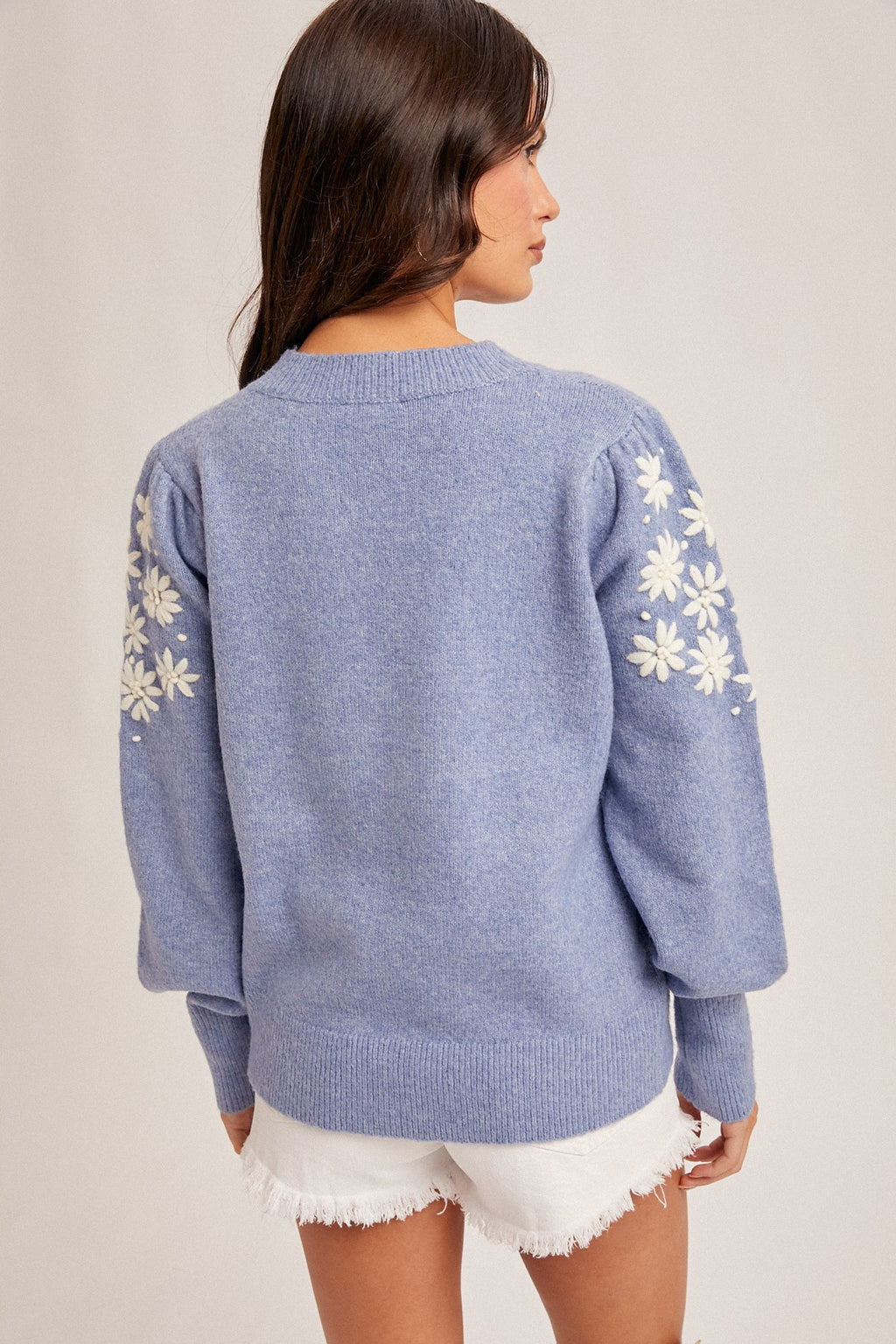 Back view of a woman wearing a frost  blue sweater with white floral embroidery on the sleeve on a plain background