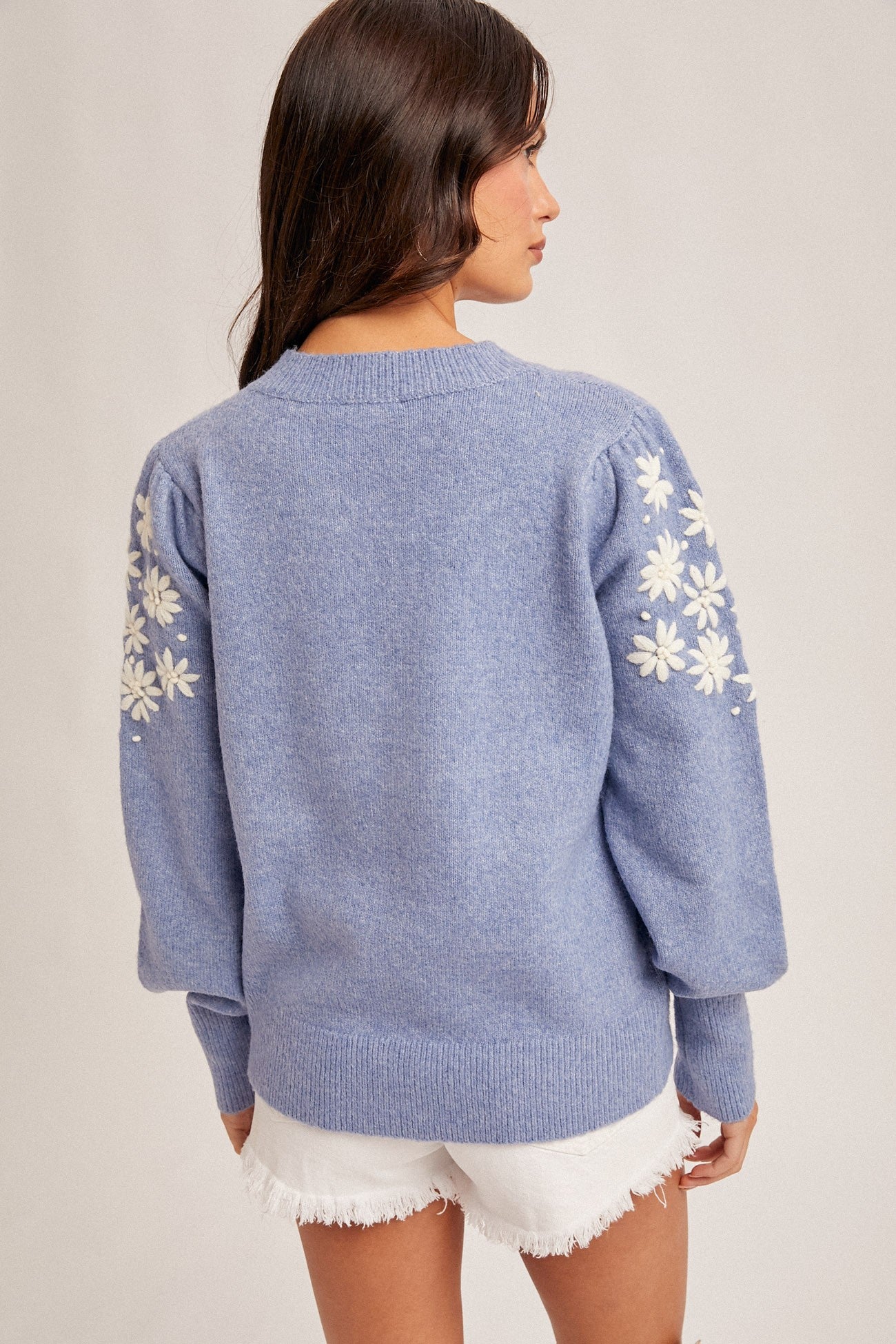 Back view of a woman wearing a frost  blue sweater with white floral embroidery on the sleeve on a plain background