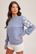 Woman wearing a frost  blue sweater with white floral embroidery on the sleeve on a plain background