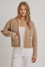 Woman wearing a open cardigan with long sleeves, front pockets and detail stitching and white pants, tan hat against a white background