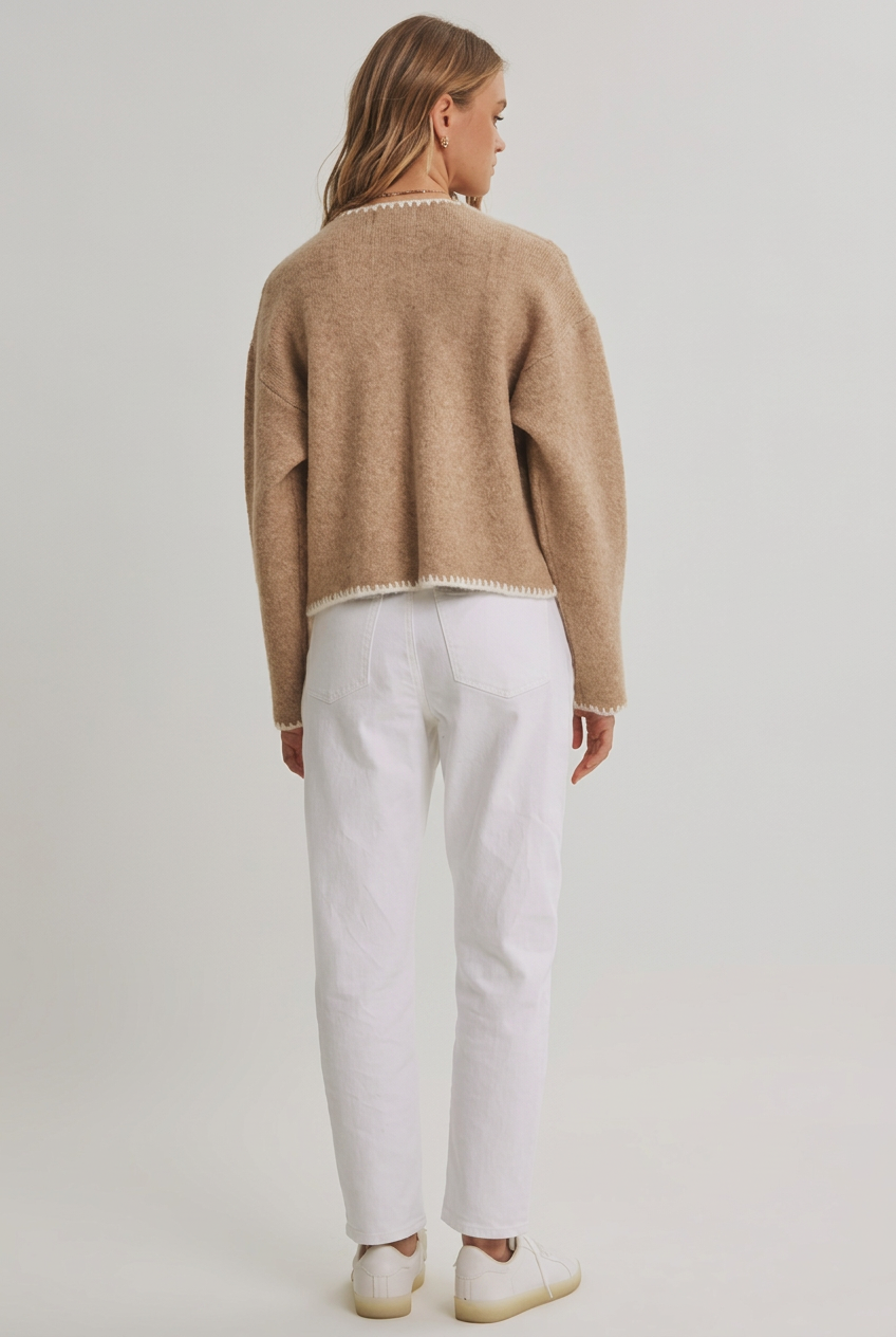 Back view of a woman wearing a open cardigan with long sleeves, front pockets and detail stitching and white pants against a white background