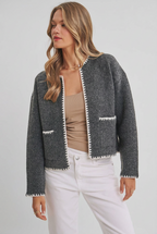 Woman wearing an open charcoal cardigan with long sleeves, front pockets, detail stitching accent with white pants against a gray background