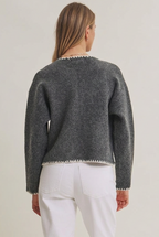 Back view of a woman wearing an open charcoal cardigan with long sleeves, front pockets, detail stitching accent with white pants against a gray background