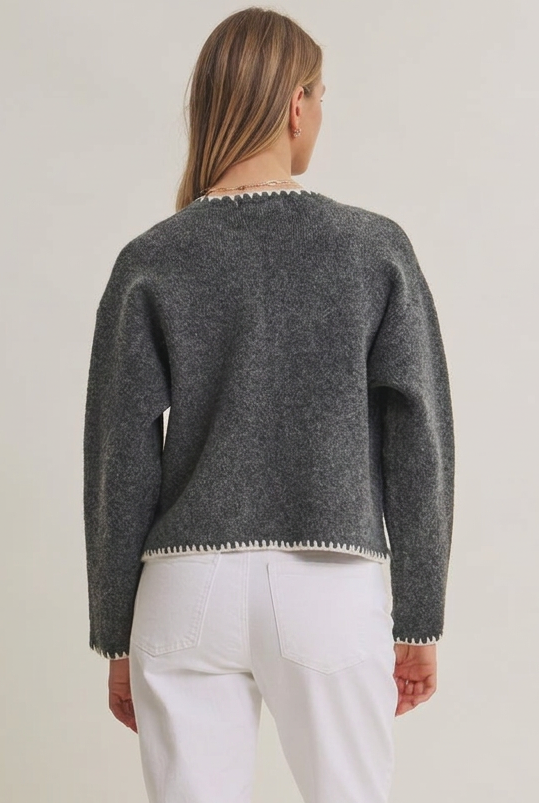 Back view of a woman wearing an open charcoal cardigan with long sleeves, front pockets, detail stitching accent with white pants against a gray background