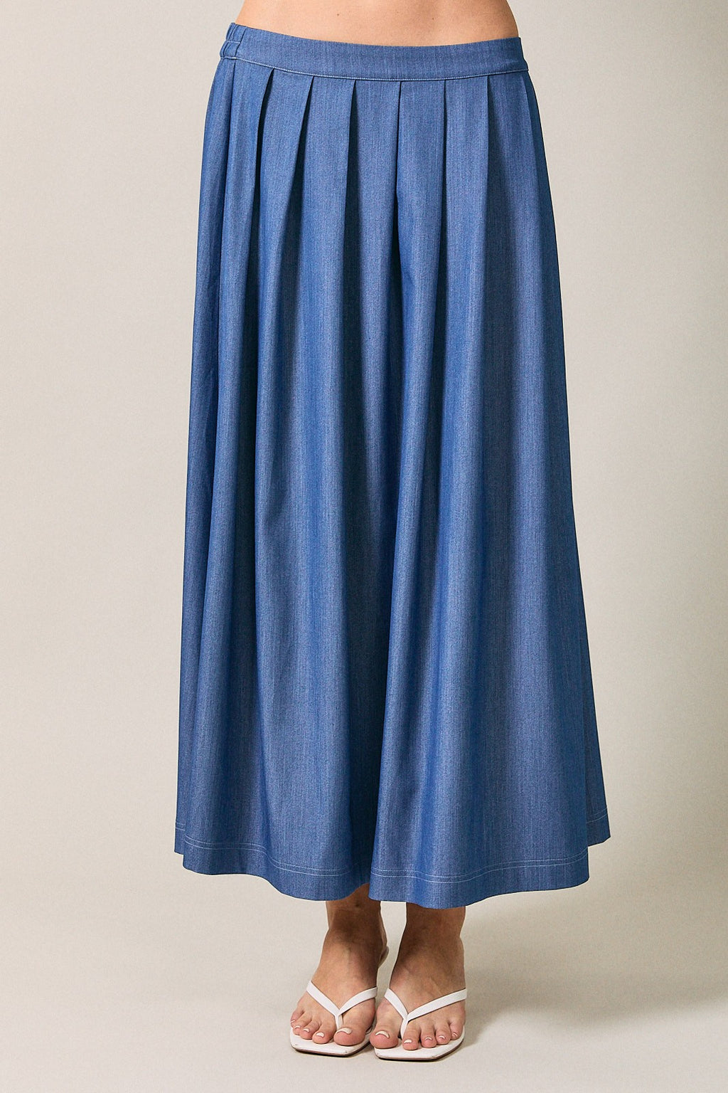 Woman wearing a denim blue pleated midi  skirt against a neutral beige studio background.