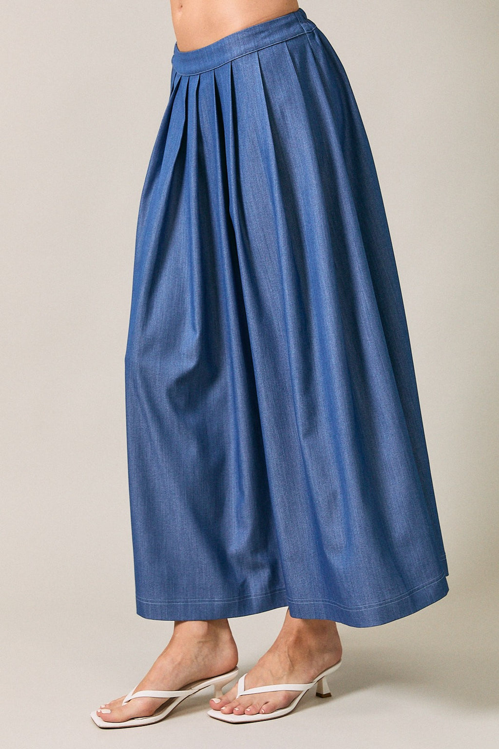 Woman wearing a denim blue pleated midi  skirt against a neutral beige studio background.