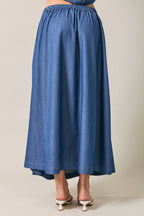 Back view of a woman wearing a denim blue pleated midi  skirt against a neutral beige studio background.