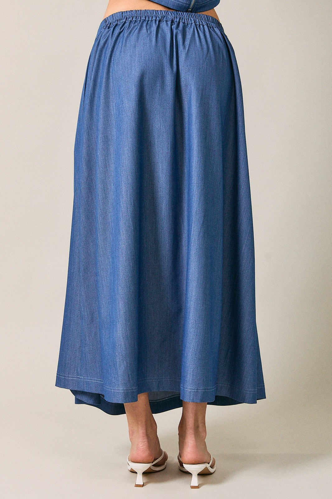 Back view of a woman wearing a denim blue pleated midi  skirt against a neutral beige studio background.