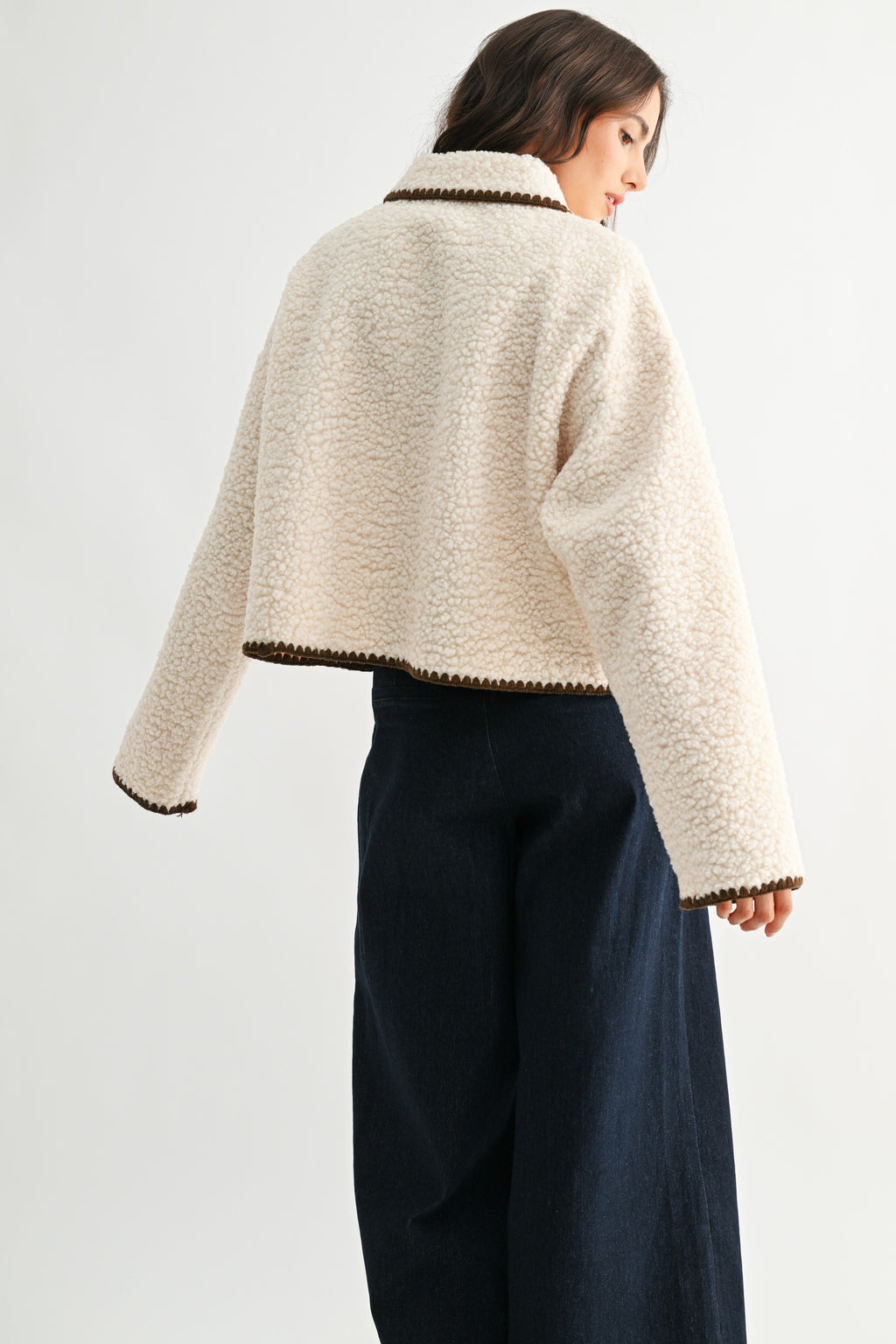 Back view of a woman wearing a cream sherpa jacket with dark brown trim and jeans on a white background 
