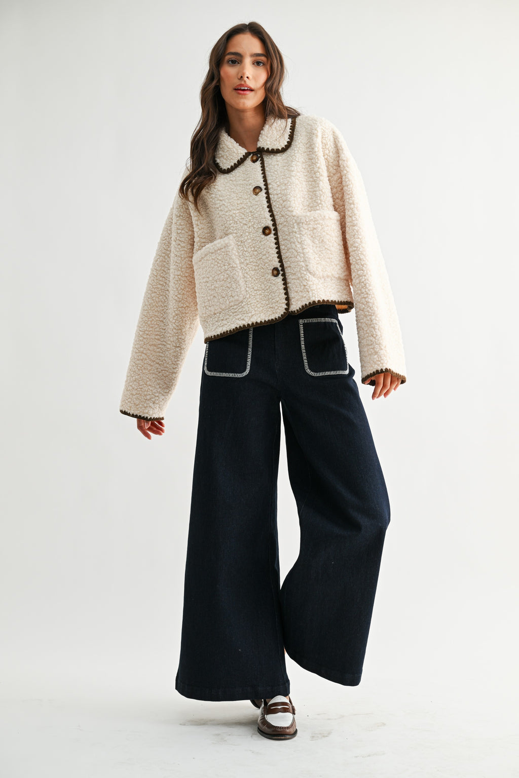 Woman wearing a cream sherpa jacket with dark brown trim and jeans on a white background 