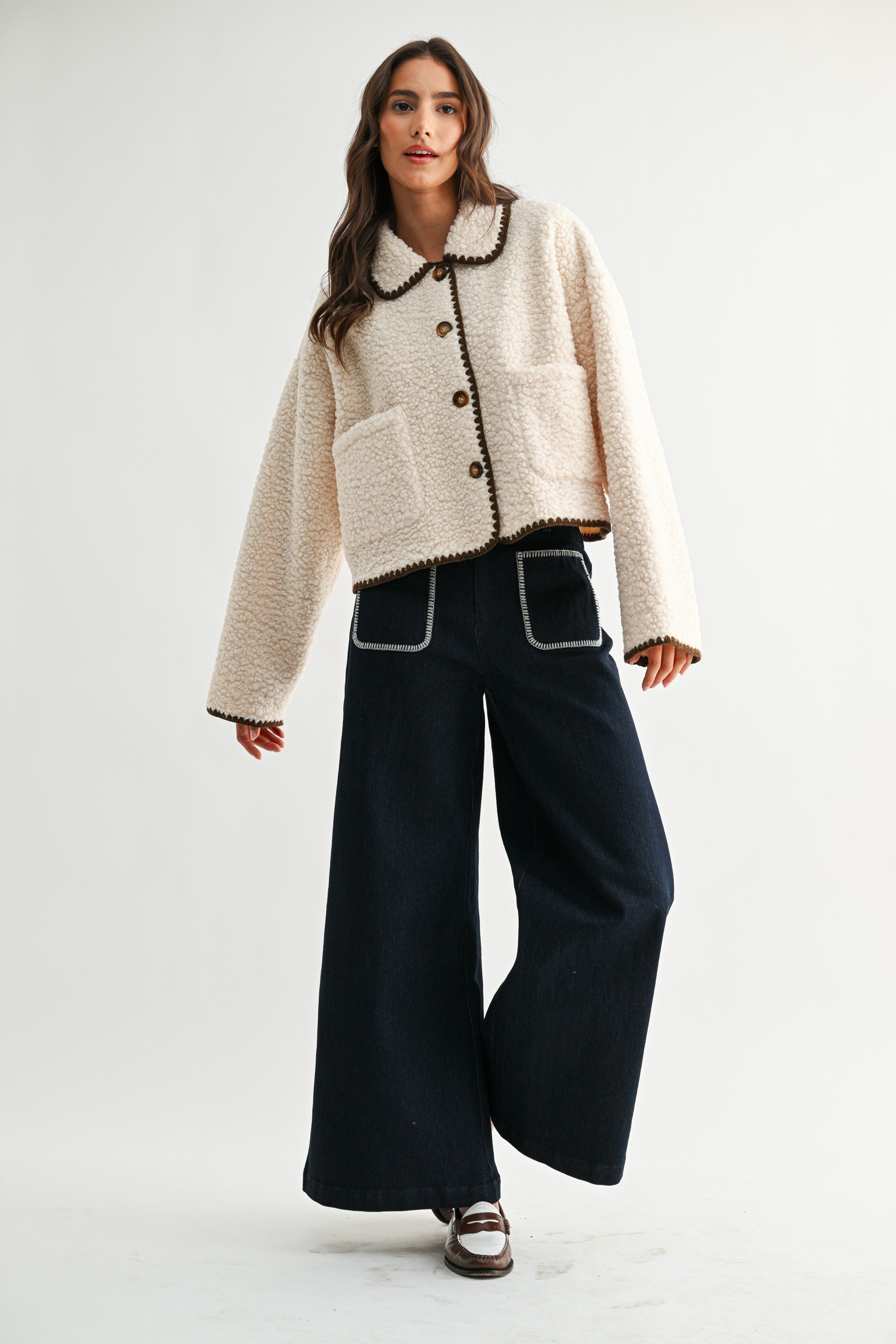 Woman wearing a cream sherpa jacket with dark brown trim and jeans on a white background 