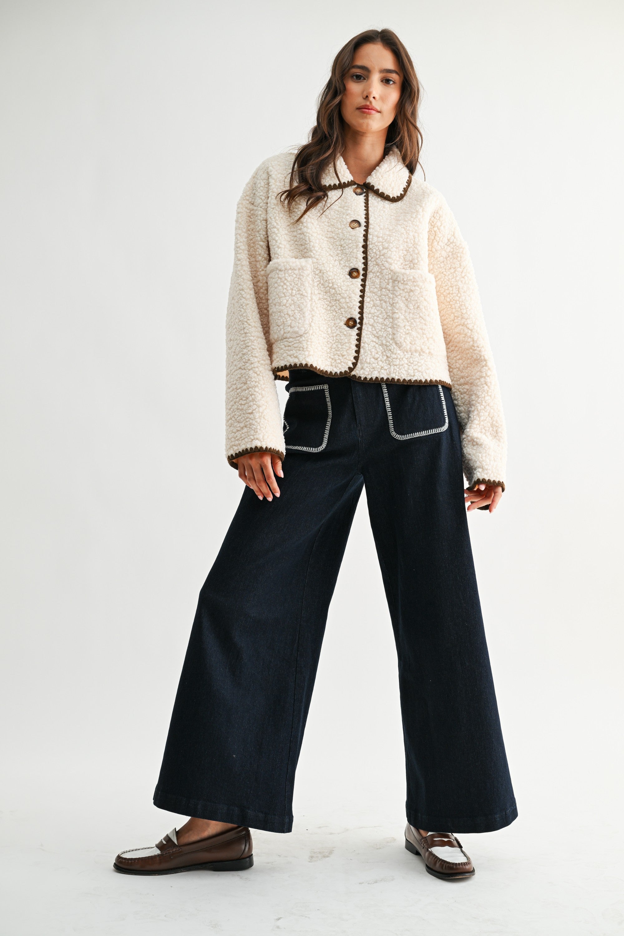 Woman wearing a cream sherpa jacket with dark brown trim and jeans on a white background 