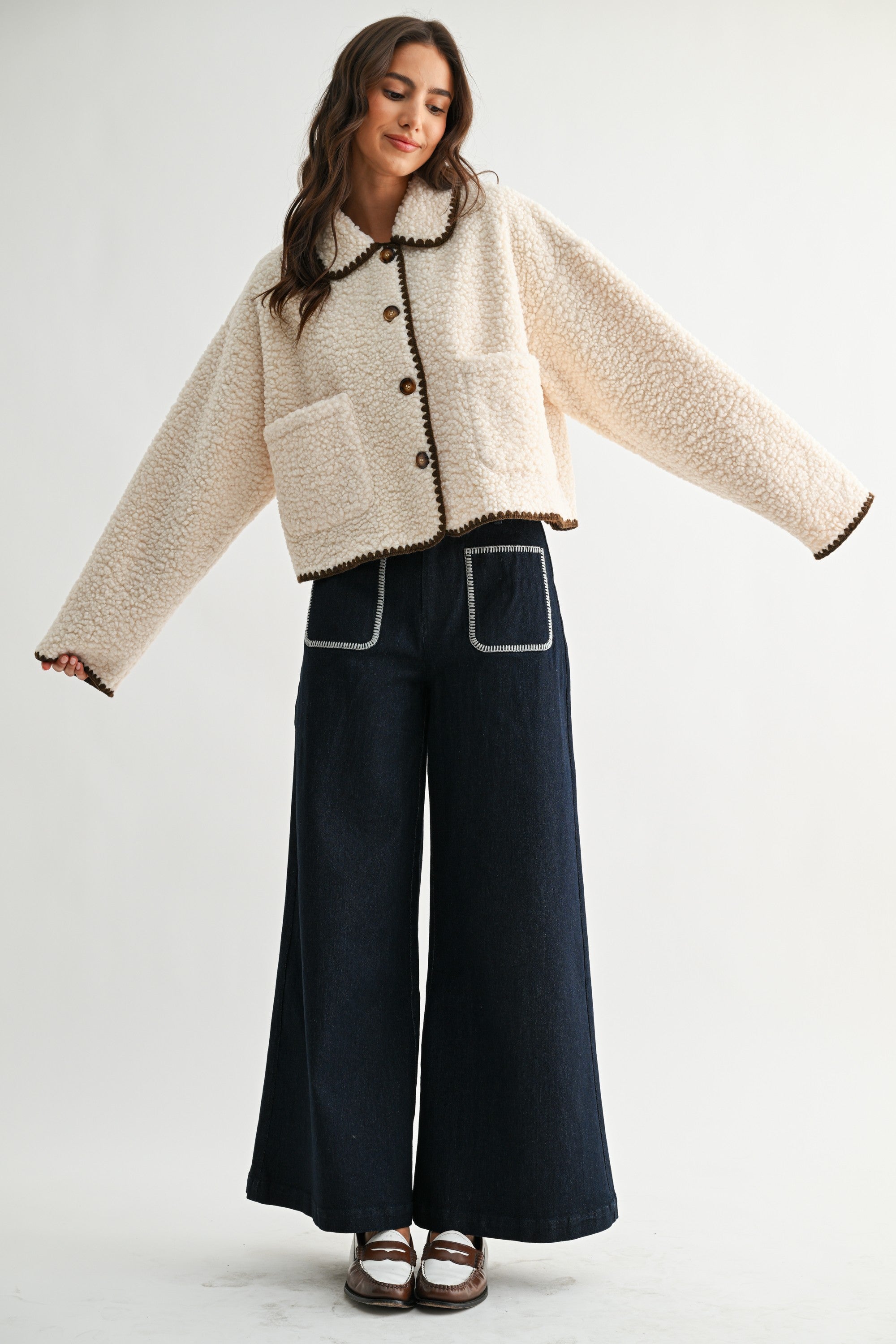 Woman wearing a cream sherpa jacket with dark brown trim and jeans on a white background 