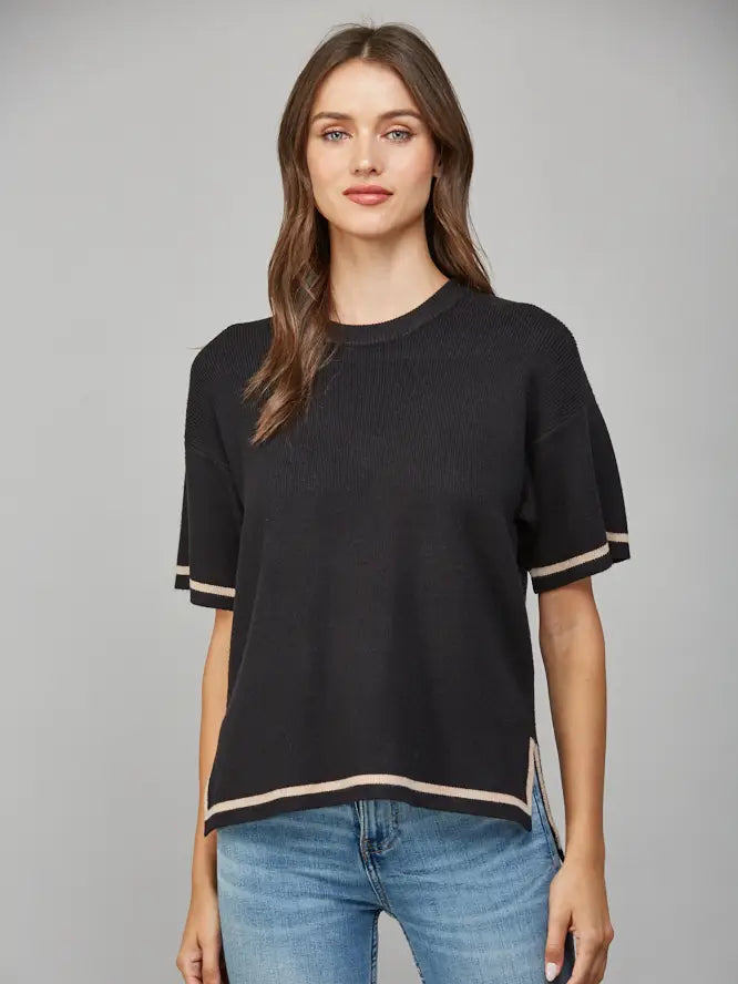 Woman wearing a black short-sleeve sweater with tan trim and blue jeans on a gray background