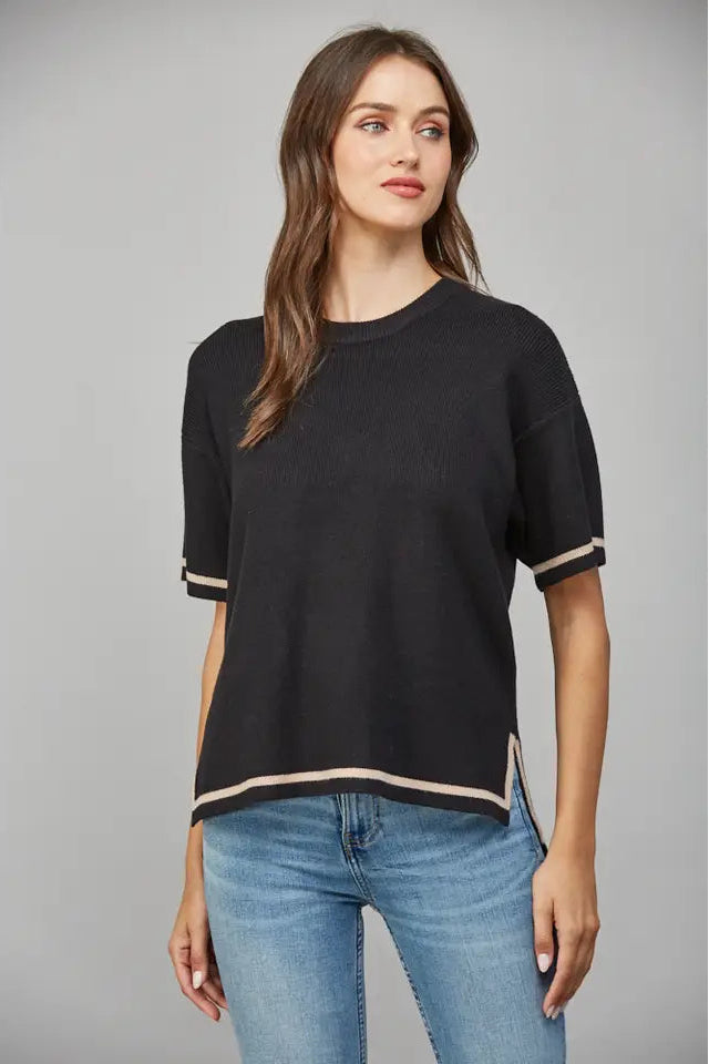 Woman wearing a black short-sleeve sweater with tan trim and blue jeans on a gray background