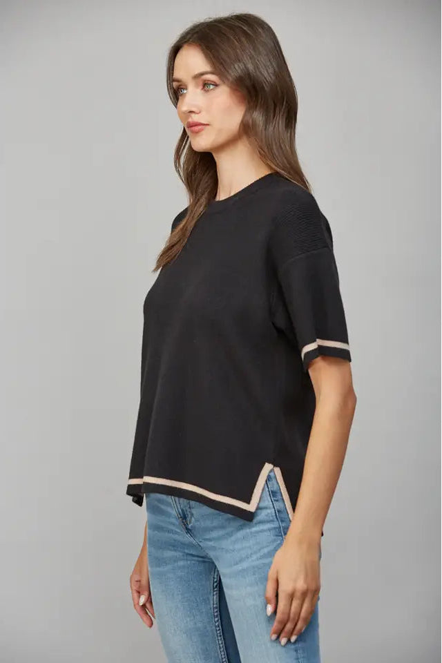 Woman wearing a black short-sleeve sweater with tan trim and blue jeans on a gray background