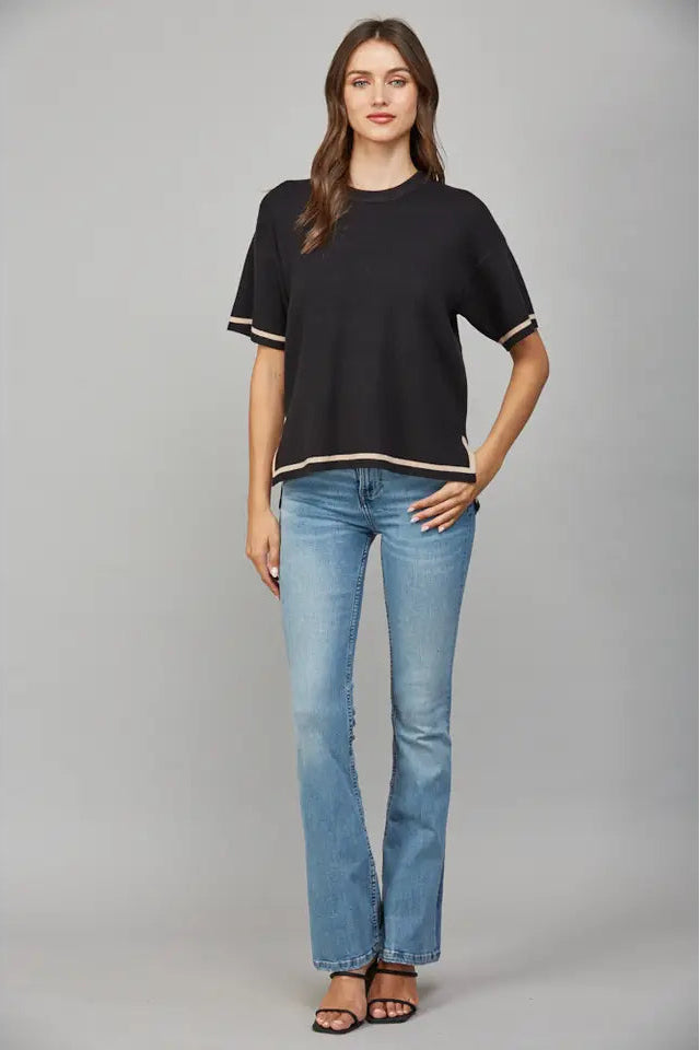 Woman wearing a black short-sleeve sweater with tan trim and blue jeans on a gray background