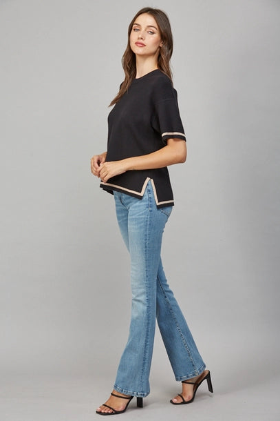 Woman wearing a black short-sleeve sweater with tan trim and blue jeans on a gray background