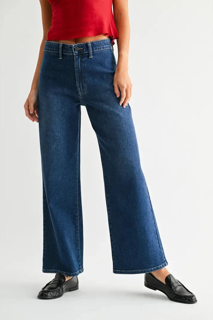A model wearing a pair of dark blue wide-leg denim jeans with no front pockets, paired with a red top and black loafers, shown from the waist down against a light gray studio background.