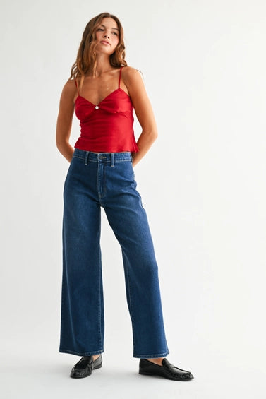 A model wearing a pair of dark blue wide-leg denim jeans with no front pockets, paired with a red top and black loafers, shown from the waist down against a light gray studio background.