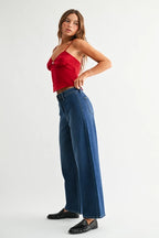 A model wearing a pair of dark blue wide-leg denim jeans with no front pockets, paired with a red top and black loafers, shown from the waist down against a light gray studio background.