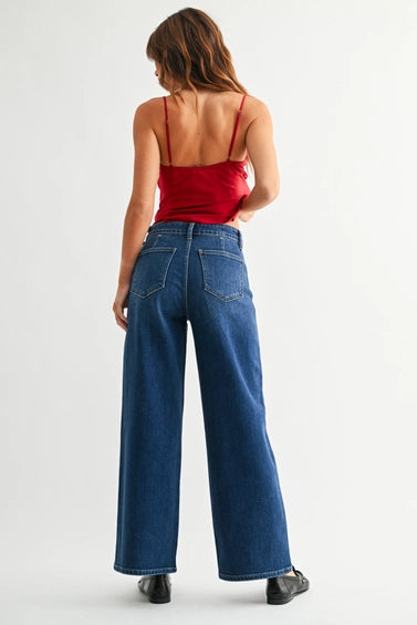 Back view of a model wearing a pair of dark blue wide-leg denim jeans with no front pockets, paired with a red top and black loafers, shown from the waist down against a light gray studio background.