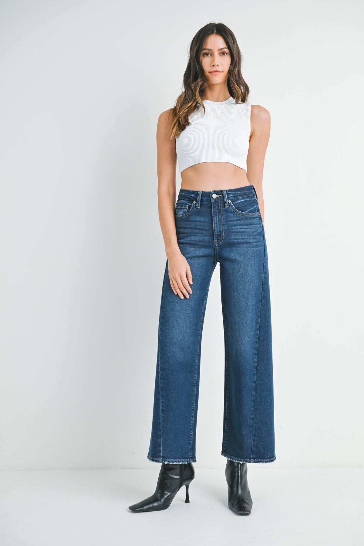 Woman wearing barrel leg dark denim jeans, white top and black high-heeled shoes on a white background
