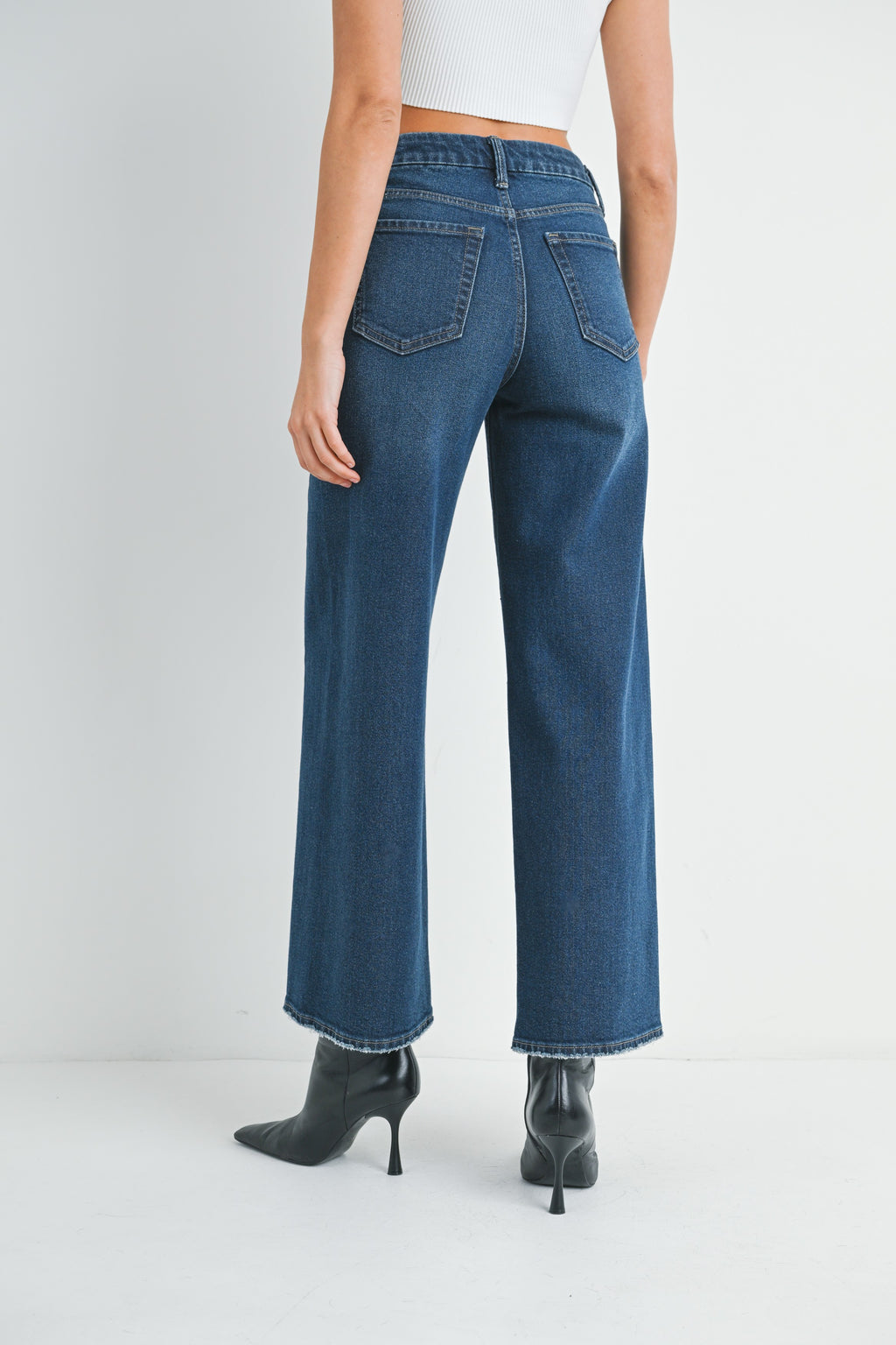 Back view of a woman wearing barrel leg dark denim jeans, white top and black high-heeled shoes on a white background