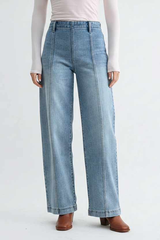 A woman modeling a pair of high-waisted, wide-leg front seam medium blue jeans with size zipper and a light pink off-the-shoulder long-sleeve top, standing against a plain light gray studio background.
